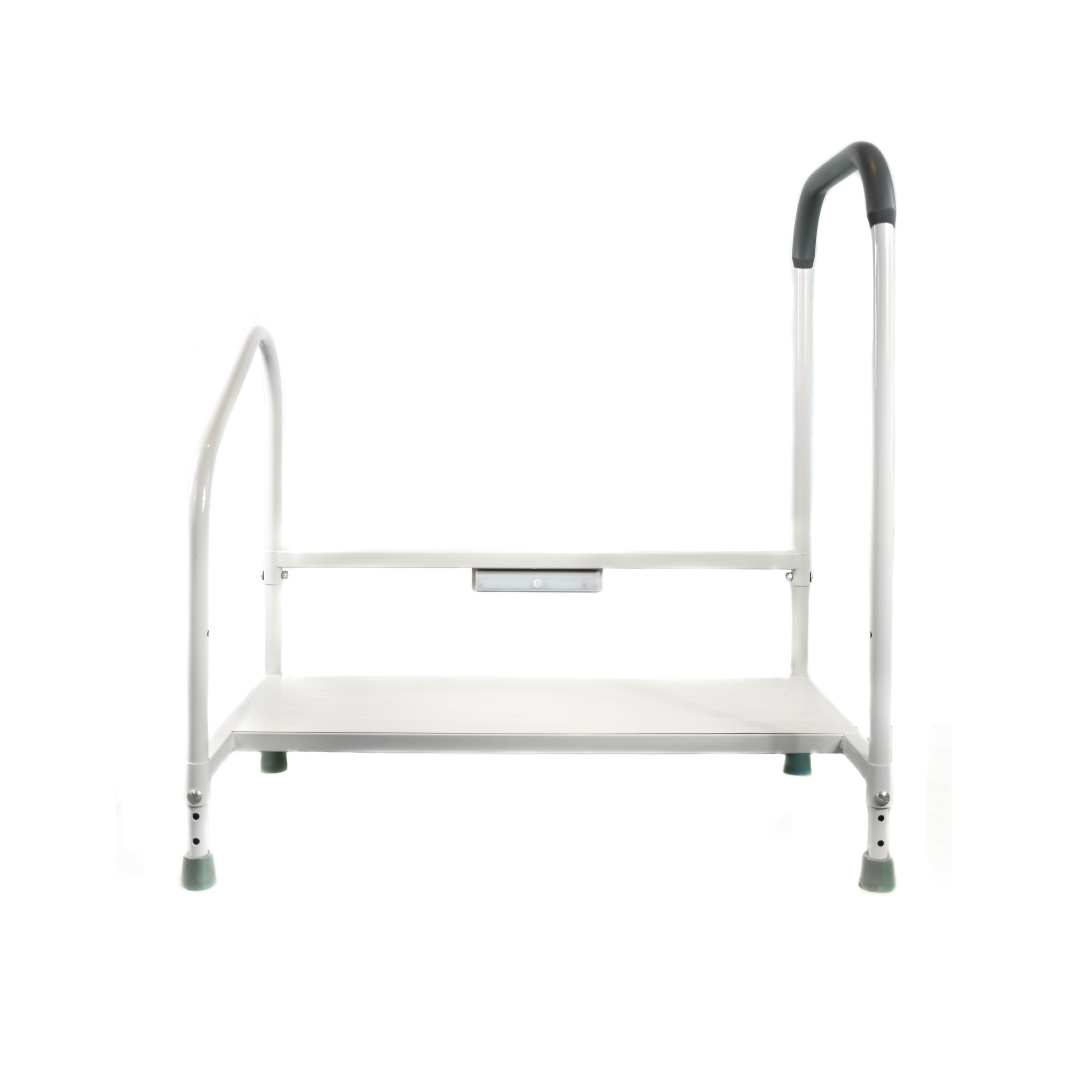 The Step2Bed XL Bedside Stepper is a white metal mobility aid with a flat platform, two support side rails, and rubber-tipped feet. A higher curved rail provides extra handhold support for added stability and fall prevention.